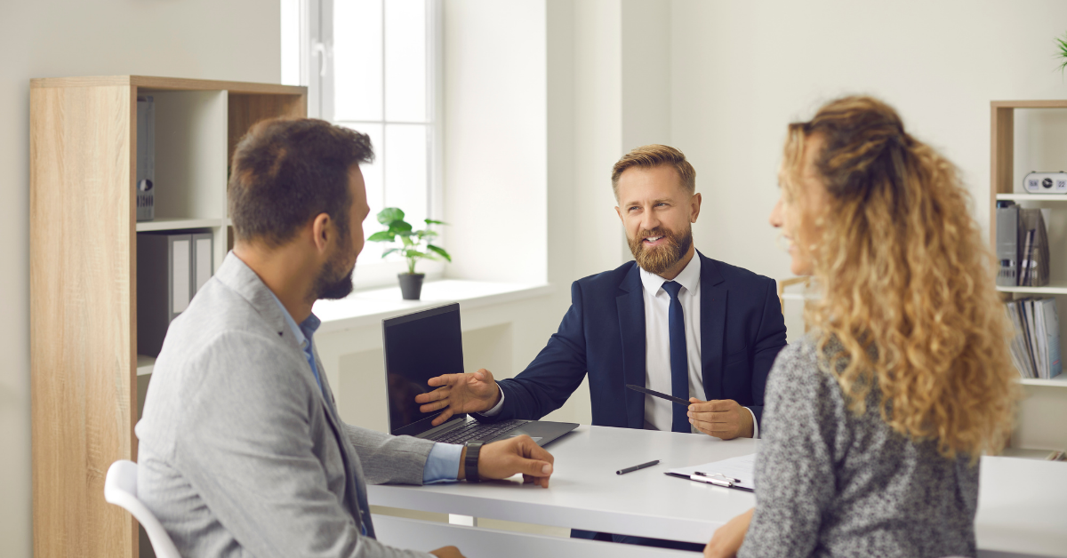Et par i møte med en smilende rådgiver i dress ved et hvitt skrivebord på et lyst kontor